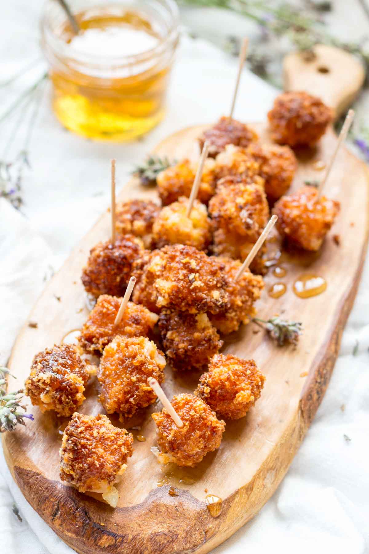 Full board of fried Manchego bites - A wooden board filled with fried Manchego cheese, lavender sprigs, and drizzled honey, with a jar of infused honey in the background.