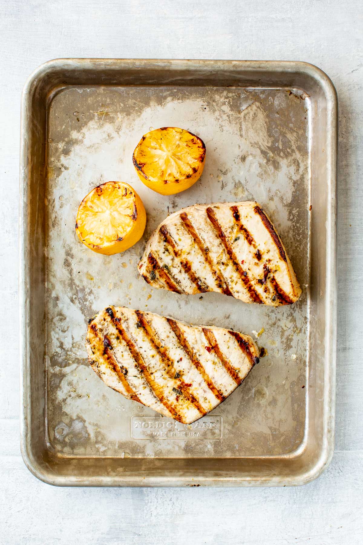 Two swordfish steaks with deep grill marks on a metal baking sheet, served alongside halved grilled lemons.