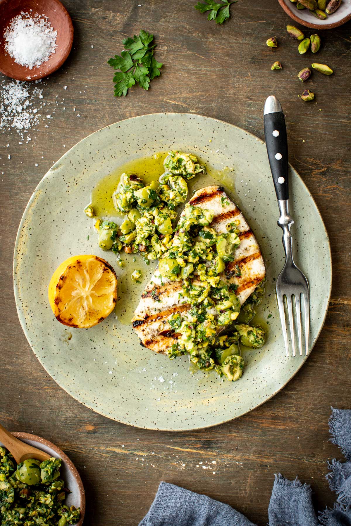 Grilled swordfish topped with olive pistachio compote on a green ceramic plate, served with a grilled lemon half and a fork.