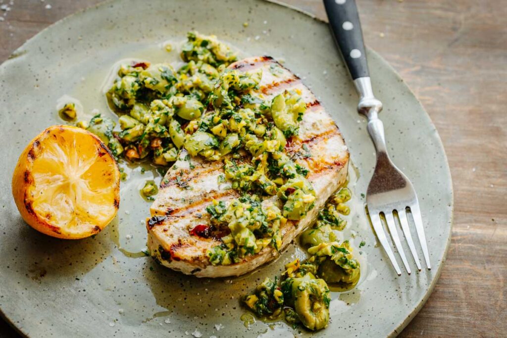 A swordfish steak topped generously with olive pistachio compote, juices pooling around, with a grilled lemon on the side.