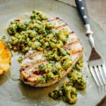 A swordfish steak topped generously with olive pistachio compote, juices pooling around, with a grilled lemon on the side.