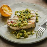 Grilled swordfish on a green plate with olive pistachio compote and charred lemon, fork placed to the right.