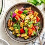 Overhead shot of plated salad and dressing - Top-down view of colorful grilled vegetable and bread salad in a dark plate, surrounded by fresh basil, pepper, and dressing.