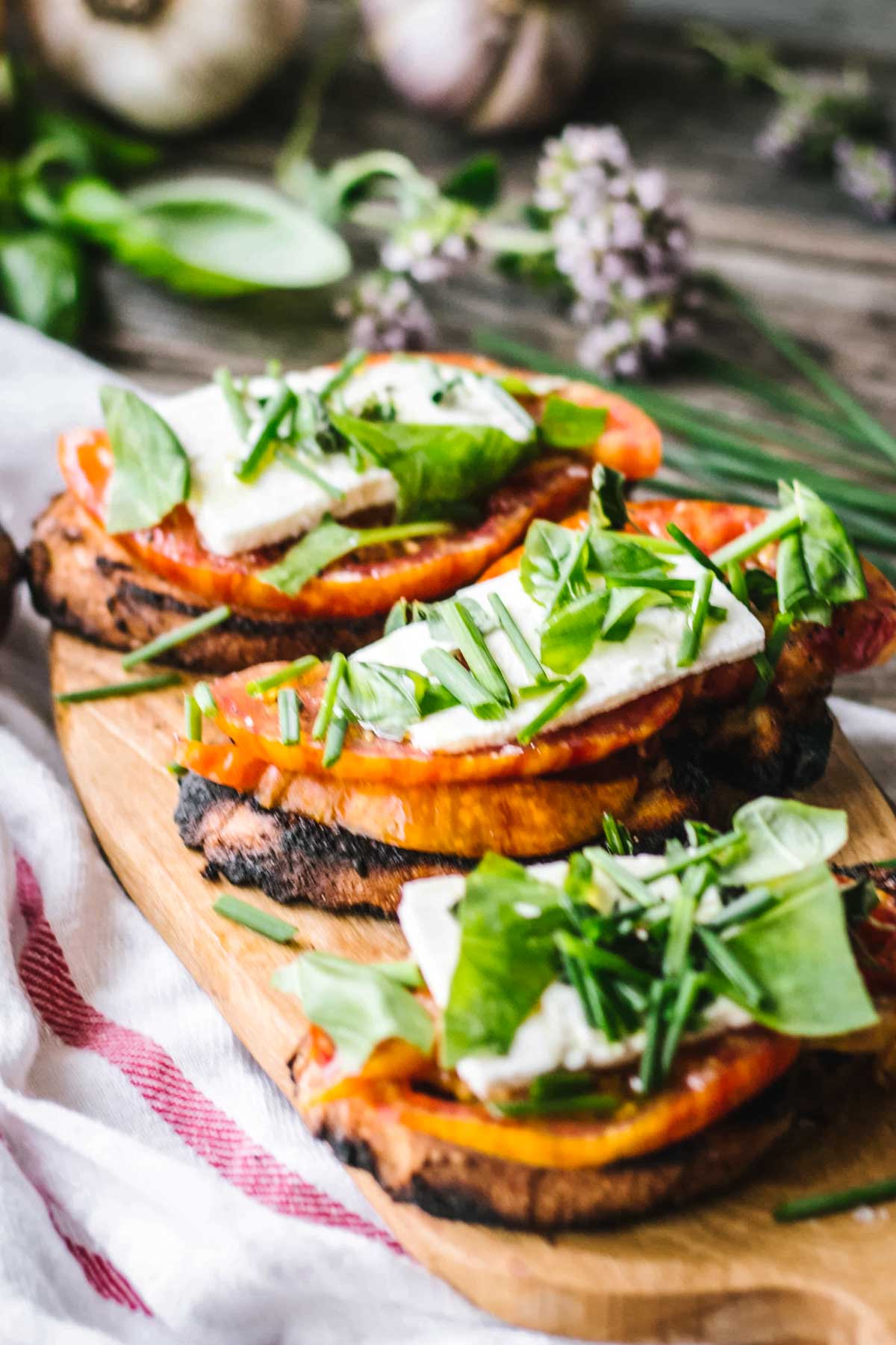Several pieces of grilled tomato and feta toasts arranged on a small wooden serving board, topped with vibrant green herbs and surrounded by garlic bulbs and flowering herbs in the background.