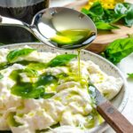 Spoon drizzling basil oil over burrata and basil leaves on a white plate.