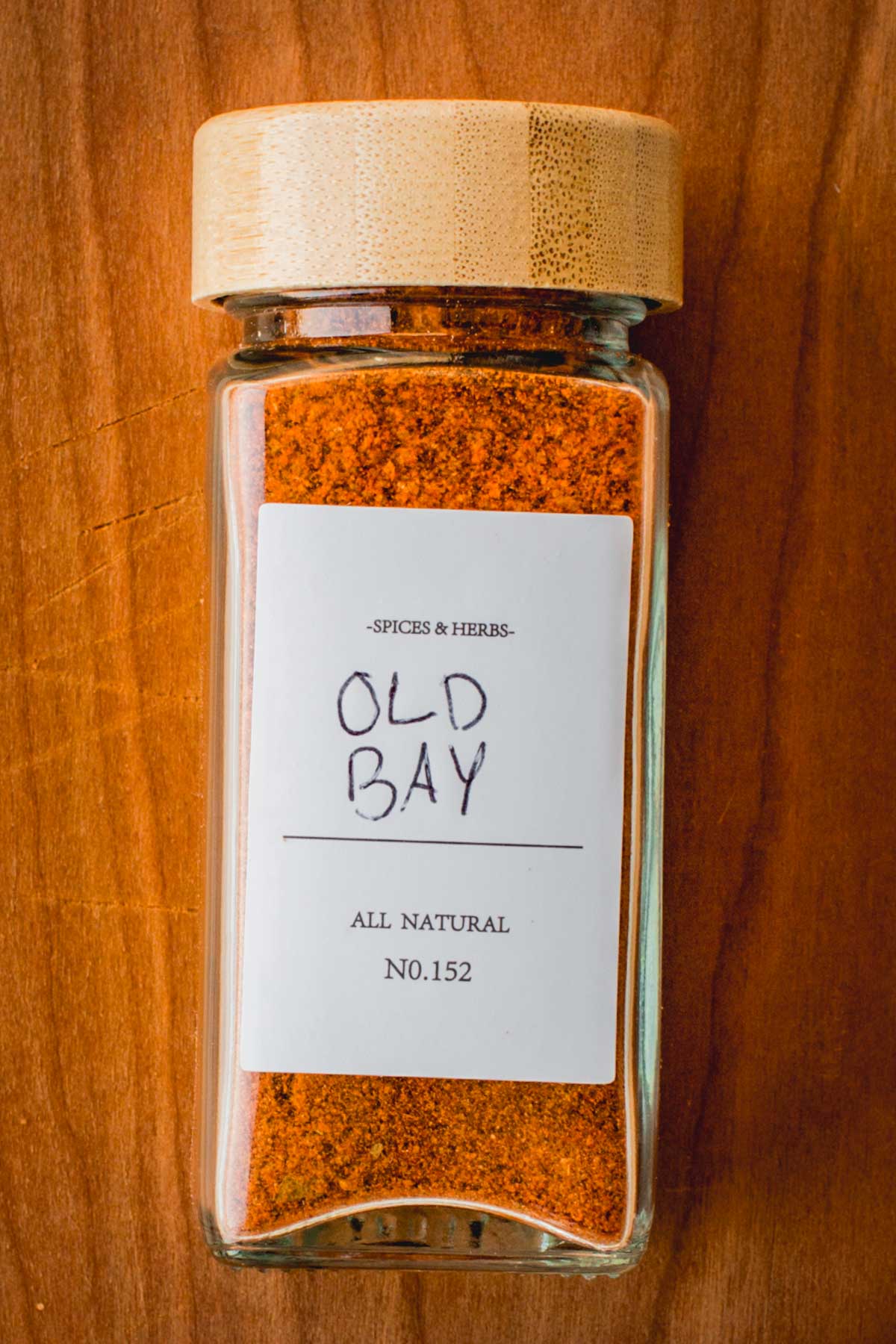 Glass spice jar labeled “Old Bay” filled with a vibrant reddish-orange homemade seasoning blend, placed on a wooden surface.
