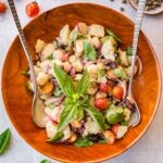 Wooden bowl of Sicilian potato salad - Insalata pantesca garnished with fresh basil sprigs, surrounded by cherry tomatoes and a small dish of salt-packed capers.