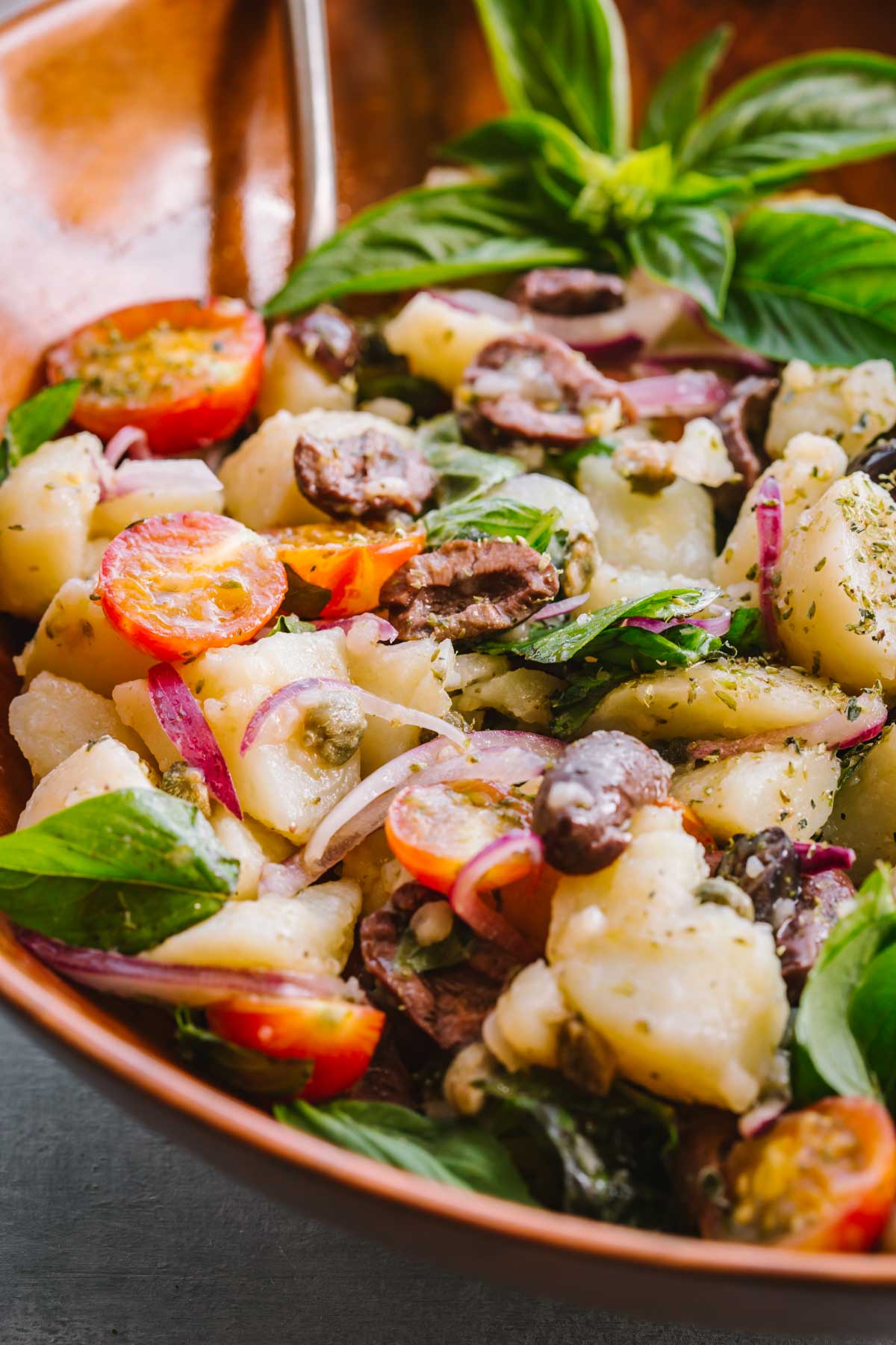 Close-up of salad in wooden bowl - Tender potato chunks, cherry tomatoes, olives, capers, red onion, and basil leaves coated in a vinegar dressing.