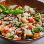 Wide bowl of insalata pantesca - A large serving of Sicilian potato salad in a wooden bowl, with bright basil leaves and glossy cherry tomatoes.