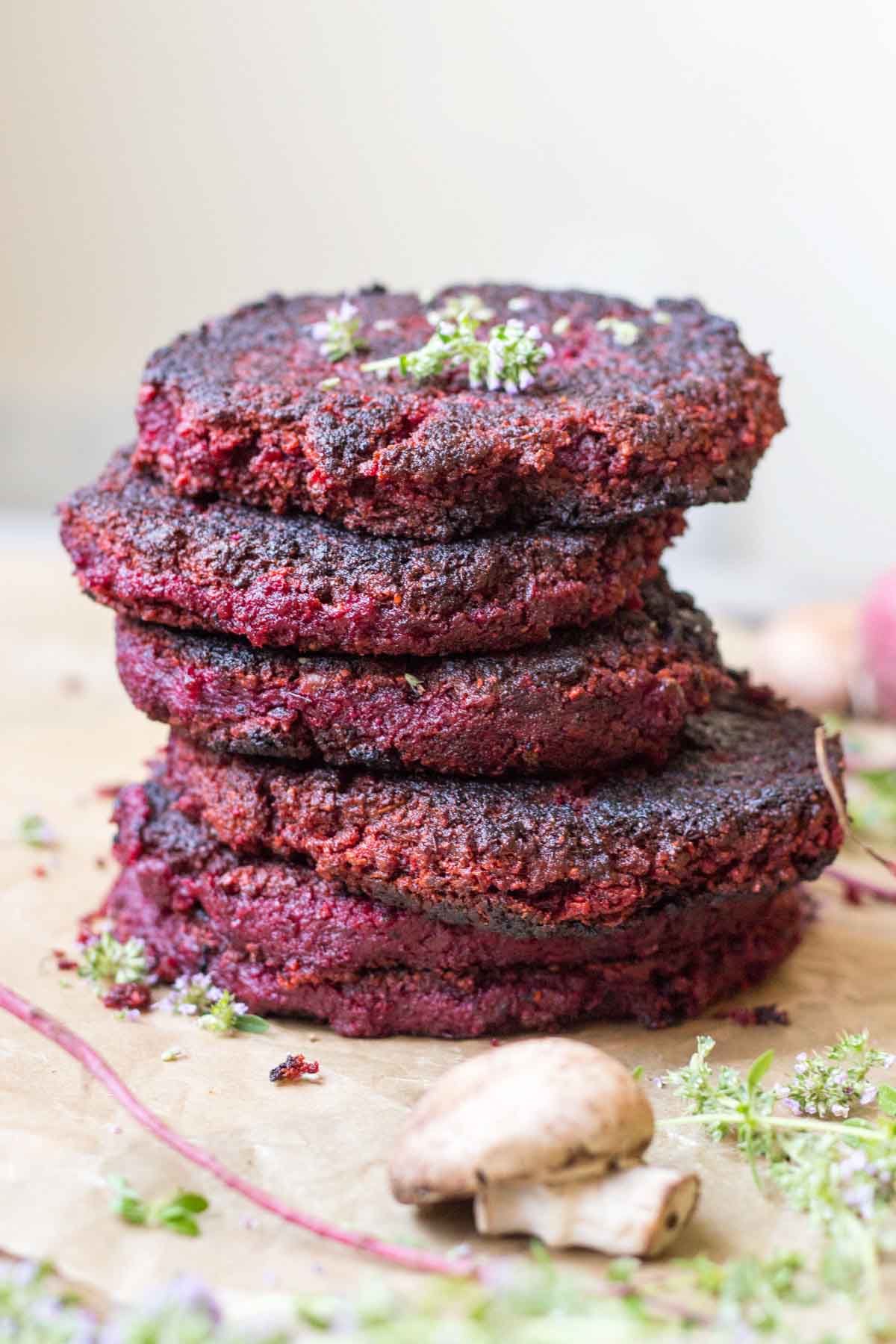 Stack of beet and lentil burgers - Four deep red lentil and beet patties stacked high on parchment paper, garnished with fresh herbs and surrounded by mushrooms and beet stems.