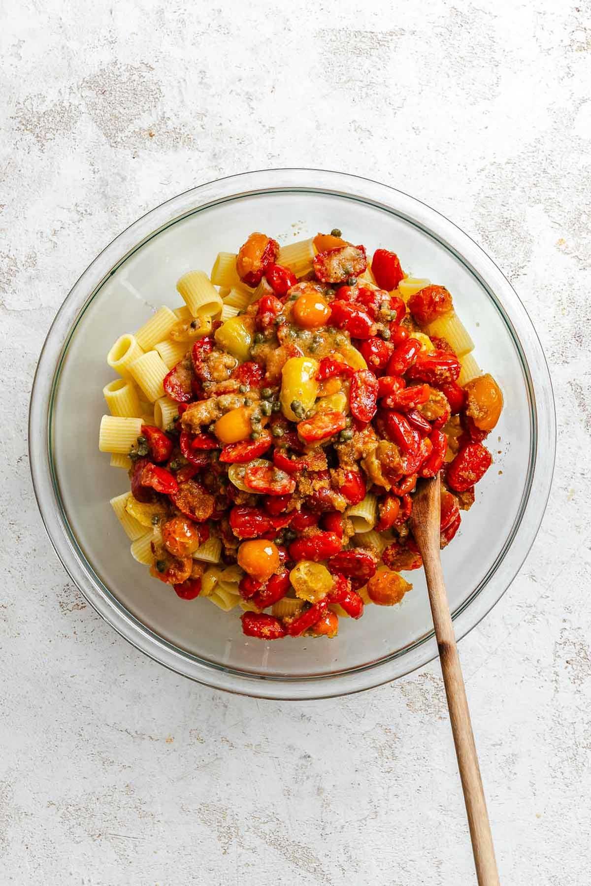Rigatoni pasta in a glass bowl topped with baked tomatoes, capers, and breadcrumbs.