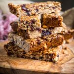 A rustic stack of chewy peanut butter and jelly granola bars on a wooden board, with visible peanuts and dried fruit, one bar with a bite taken out.