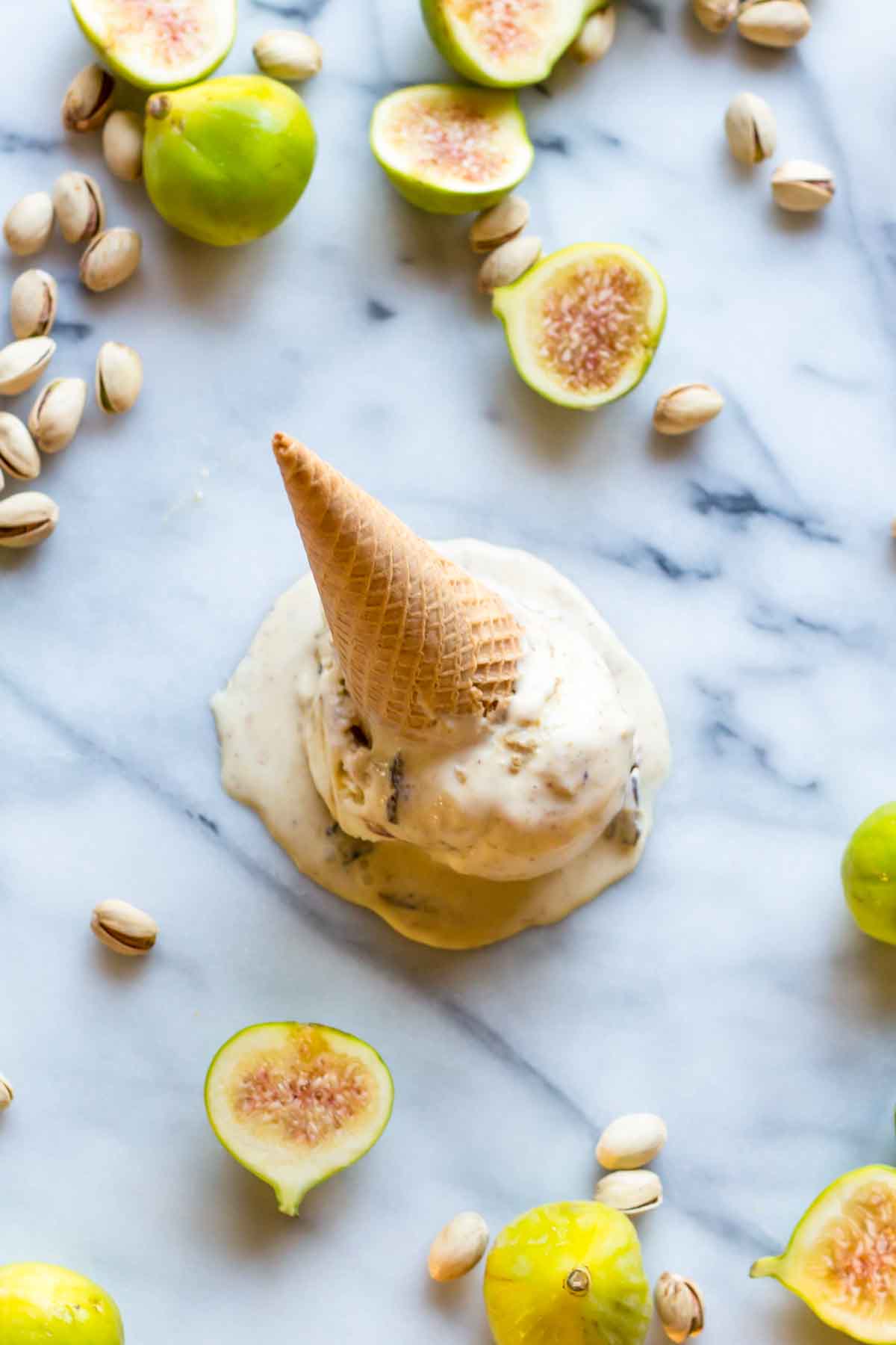 A melting scoop of ricotta, pistachio and fig gelato topped with an upside-down cone, photographed from above with figs and pistachios scattered around.