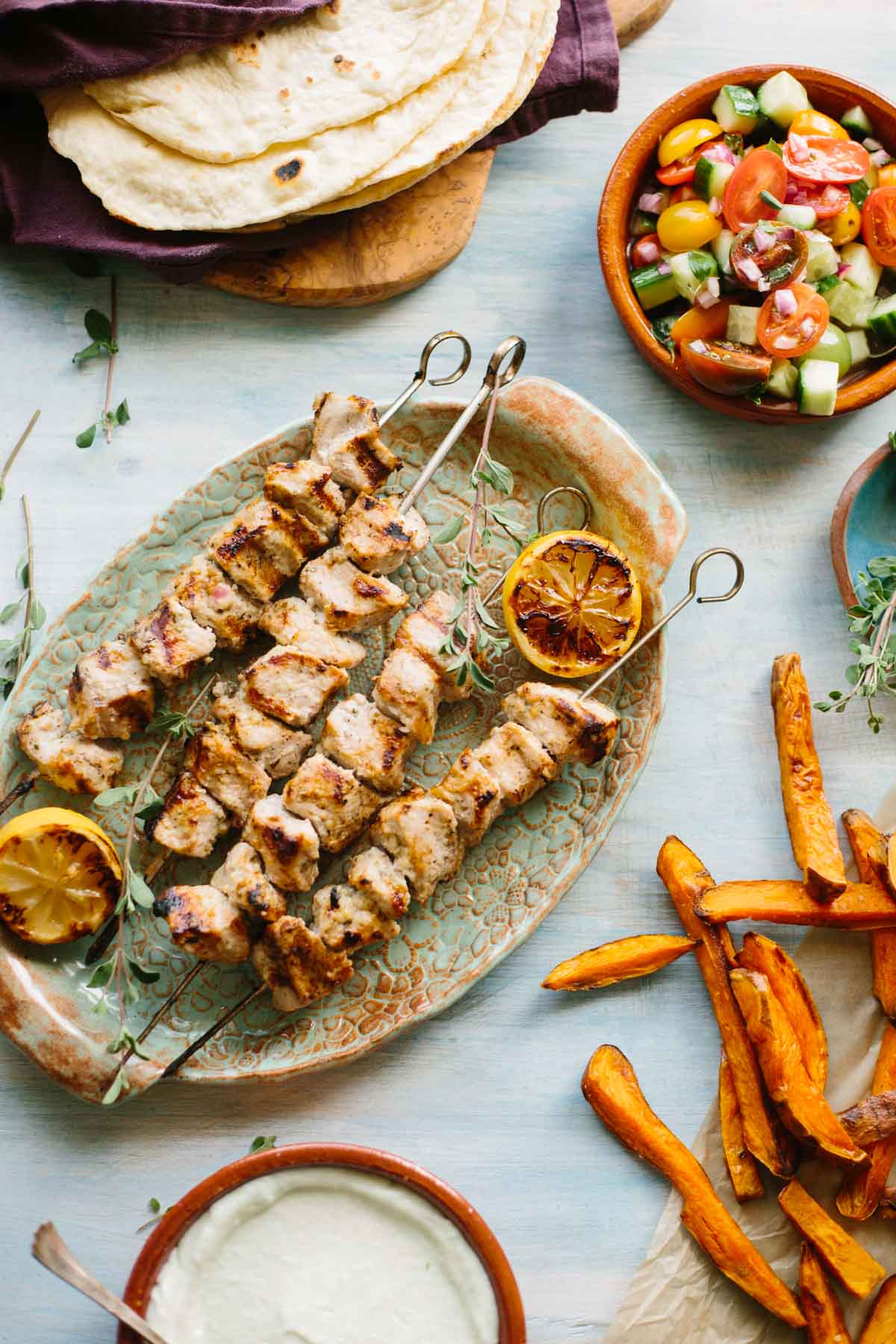 Grilled pork souvlaki skewers on a patterned platter with charred lemon halves, served with sweet potato fries, a cucumber tomato salad, warm pita bread, and a bowl of tzatziki sauce.