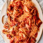 Large oval platter filled with spaghetti and crab pieces in a rich red tomato sauce, served with a fork and spoon on the side, resting on newspaper.