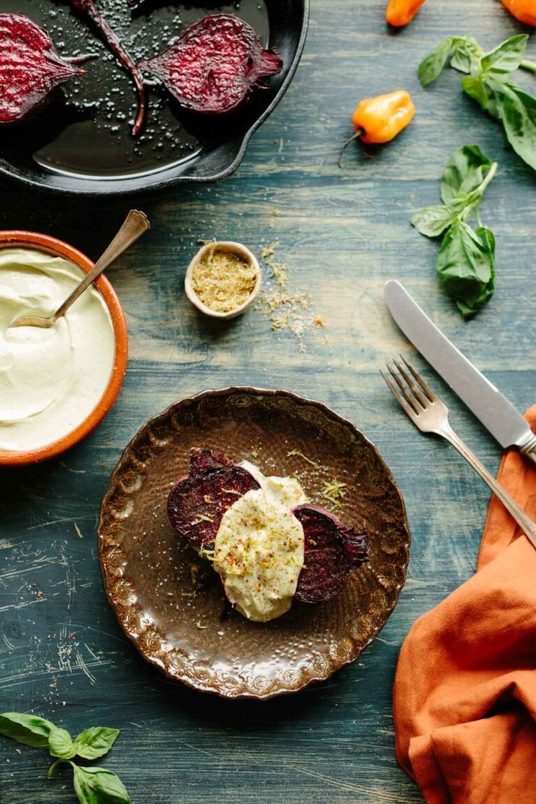 Rustic plate with roasted beets topped with avocado crema, surrounded by habaneros, fresh basil, and bowls of crema and chile lime salt on a weathered blue surface.