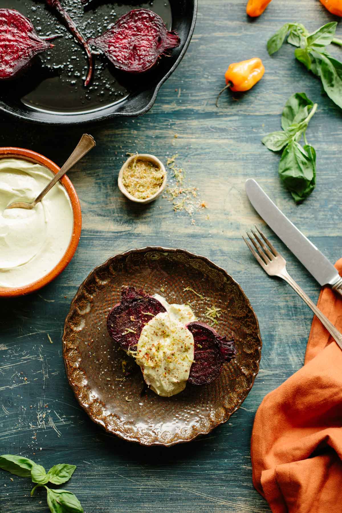 Rustic plate with roasted beets topped with avocado crema, surrounded by habaneros, fresh basil, and bowls of crema and chile lime salt on a weathered blue surface.