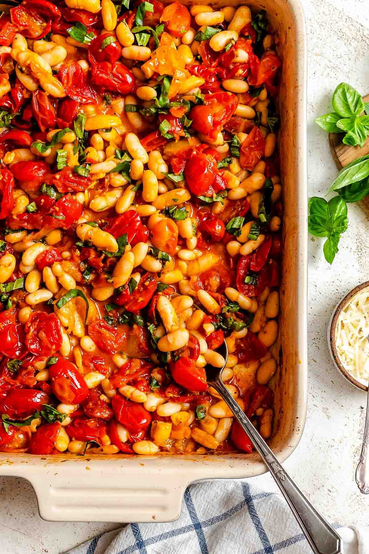 A close-up of a baking dish filled with roasted tomatoes, cannellini beans, and basil, with a spoon tucked inside for serving.