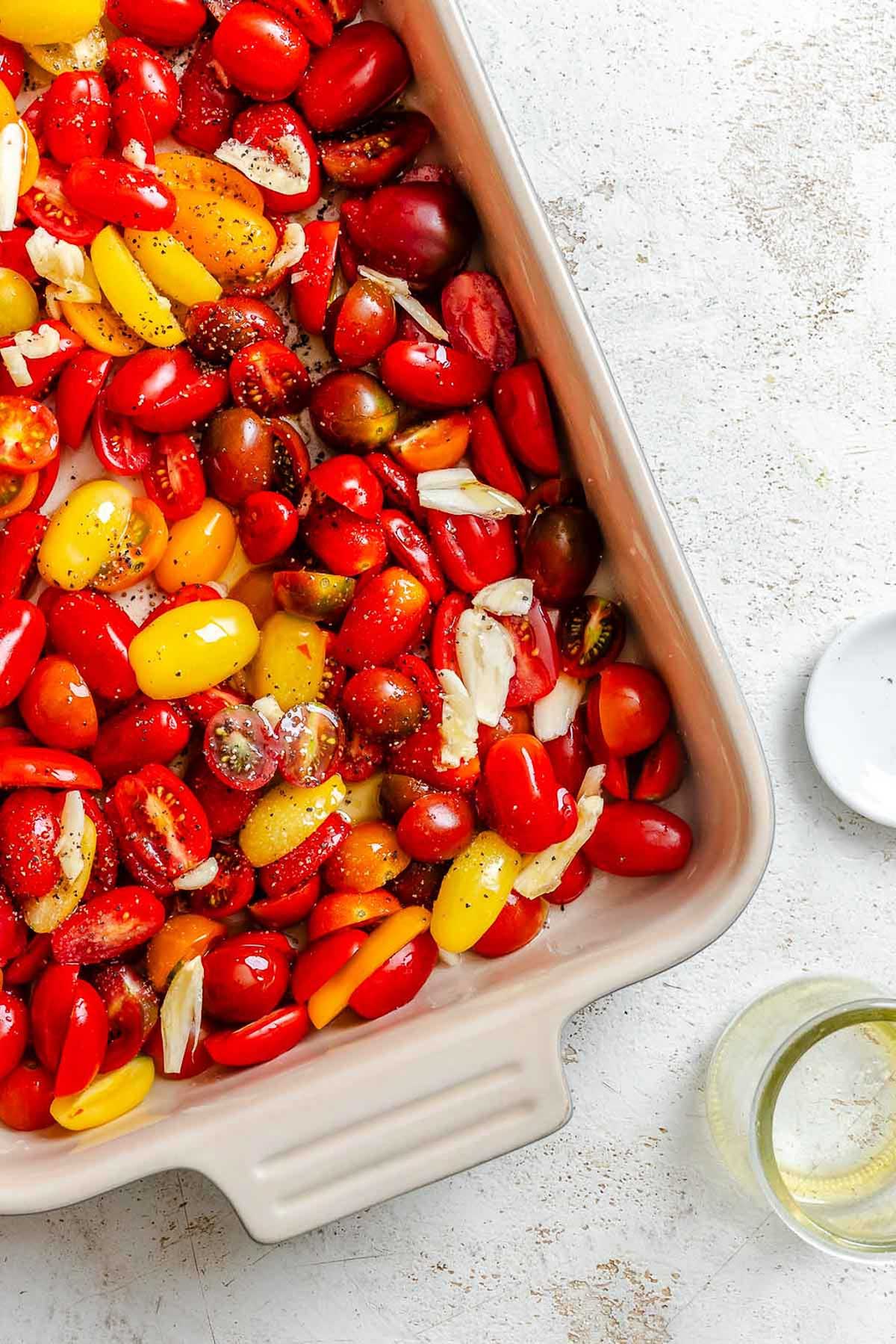 A ceramic baking dish filled with halved cherry and grape tomatoes in red, yellow, and orange, mixed with sliced garlic cloves, black pepper, and a drizzle of olive oil, ready for roasting.