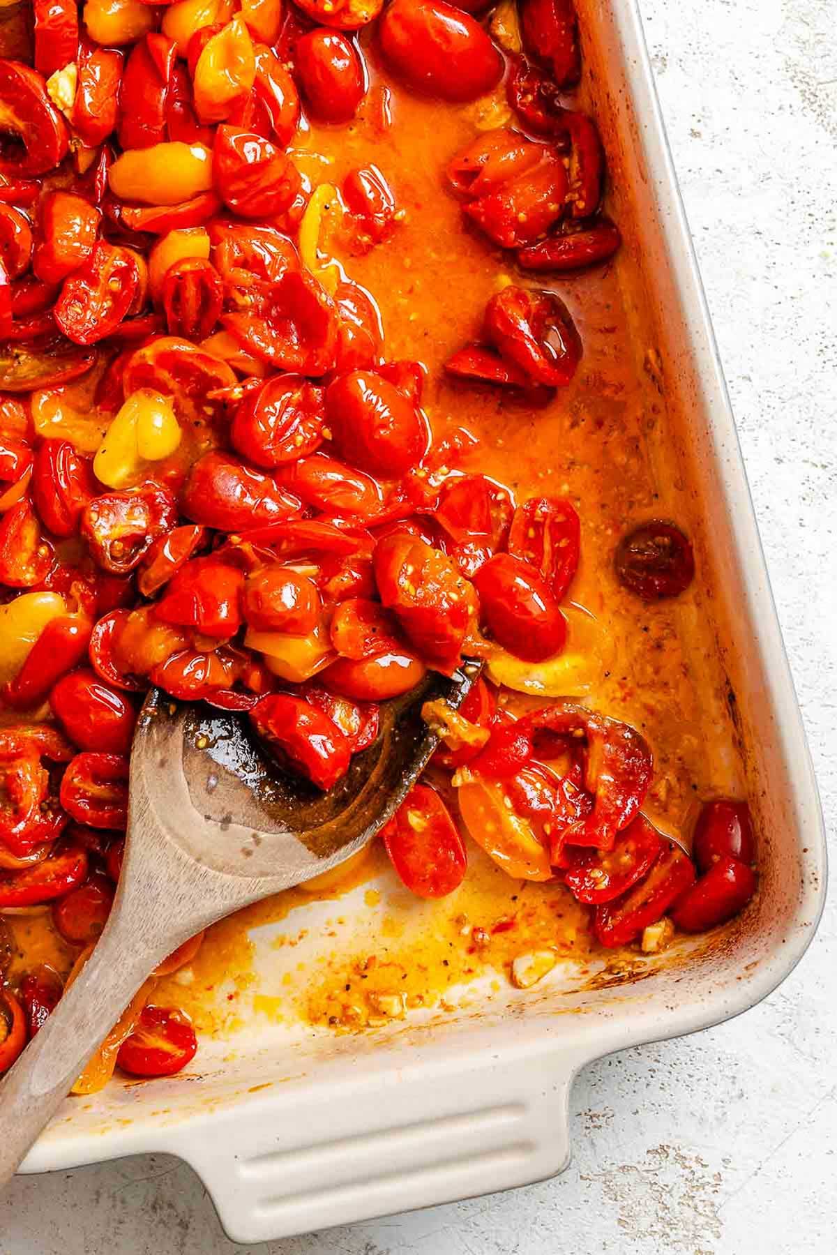 Roasted cherry tomatoes in a baking dish, softened and blistered with juices released, being stirred with a wooden spoon.