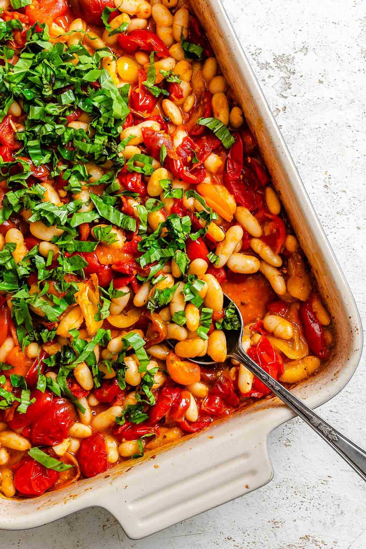 Roasted tomatoes and beans topped with a generous layer of freshly chopped basil in a baking dish, ready to be stirred together.