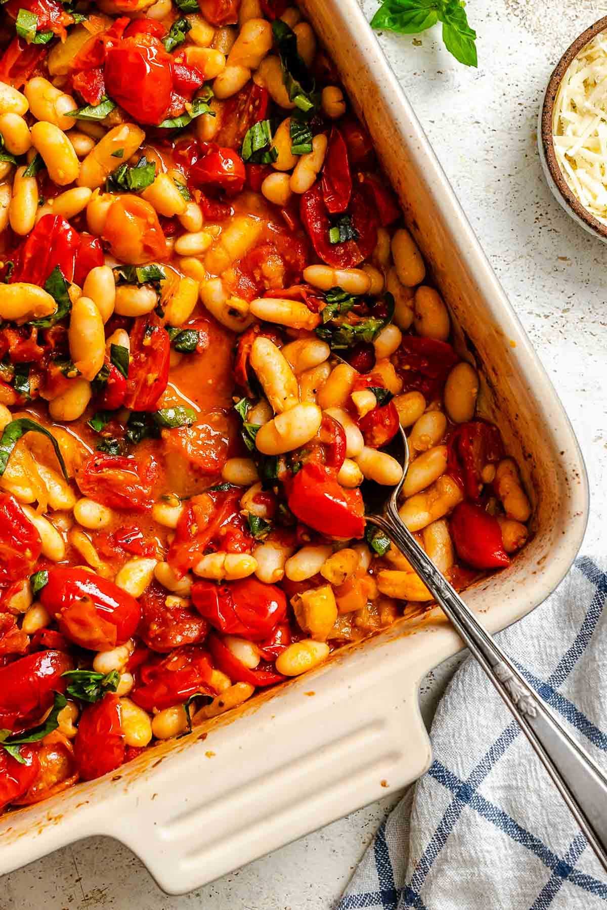 The finished roasted tomatoes with cannellini beans and basil in a baking dish, a serving spoon resting inside.