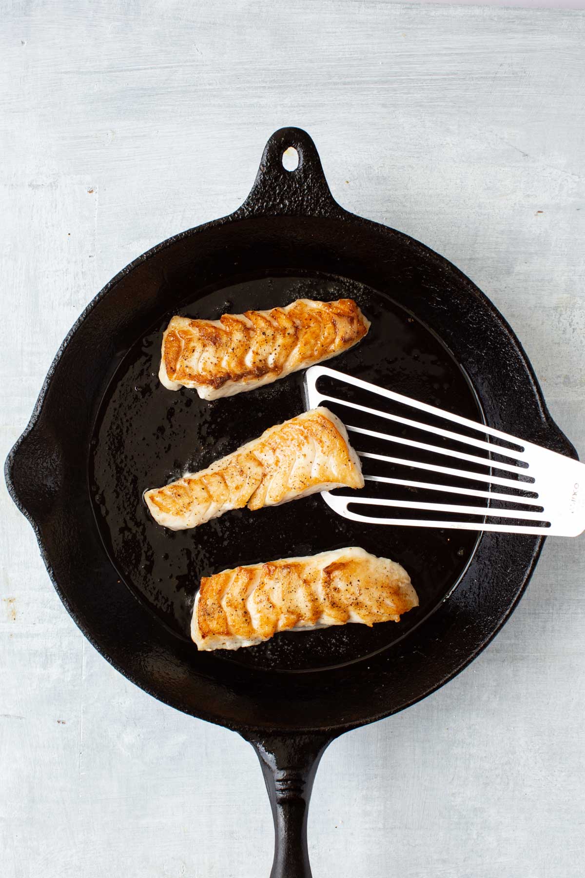 A fish spatula flipping a golden brown tilefish filet in a hot cast iron skillet.