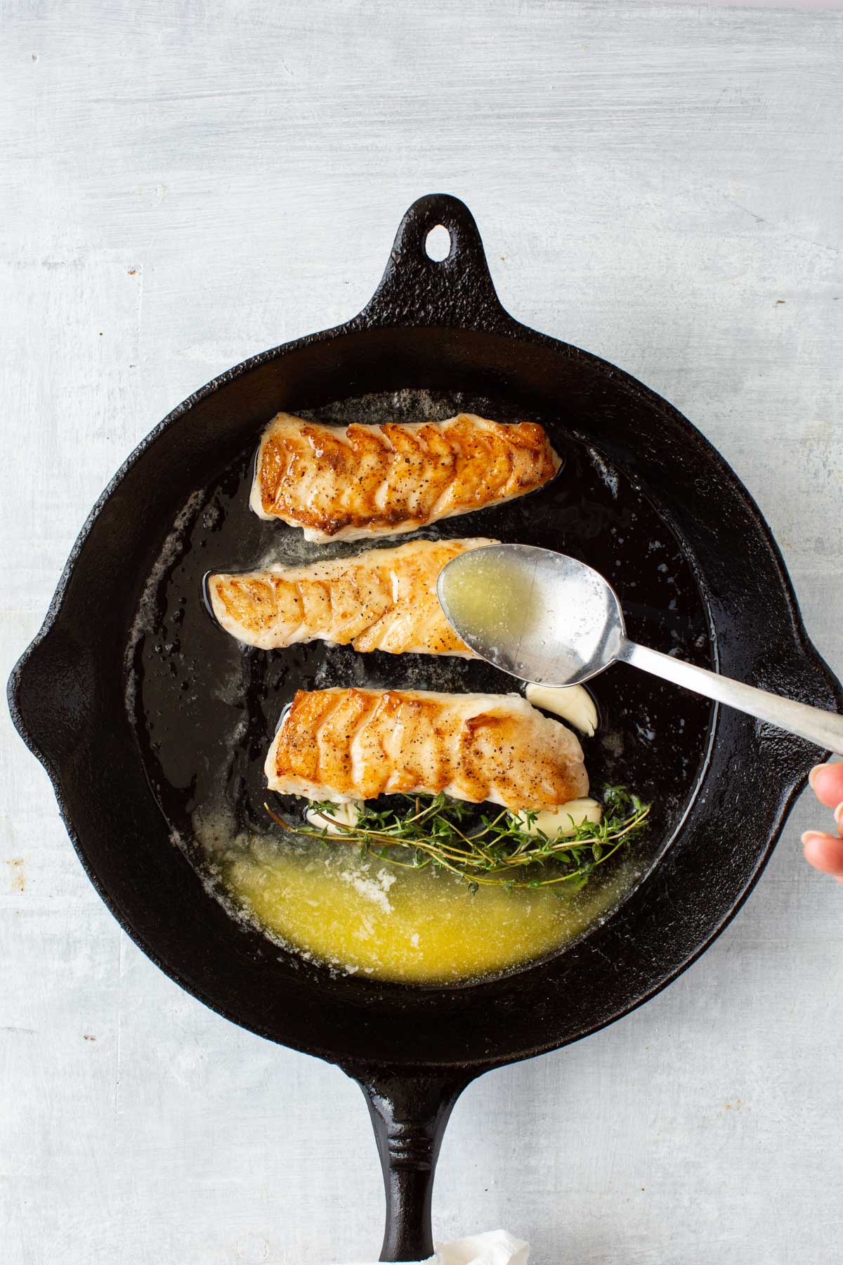 A spoon basting hot garlic-thyme butter over golden brown tilefish fillets in a skillet.