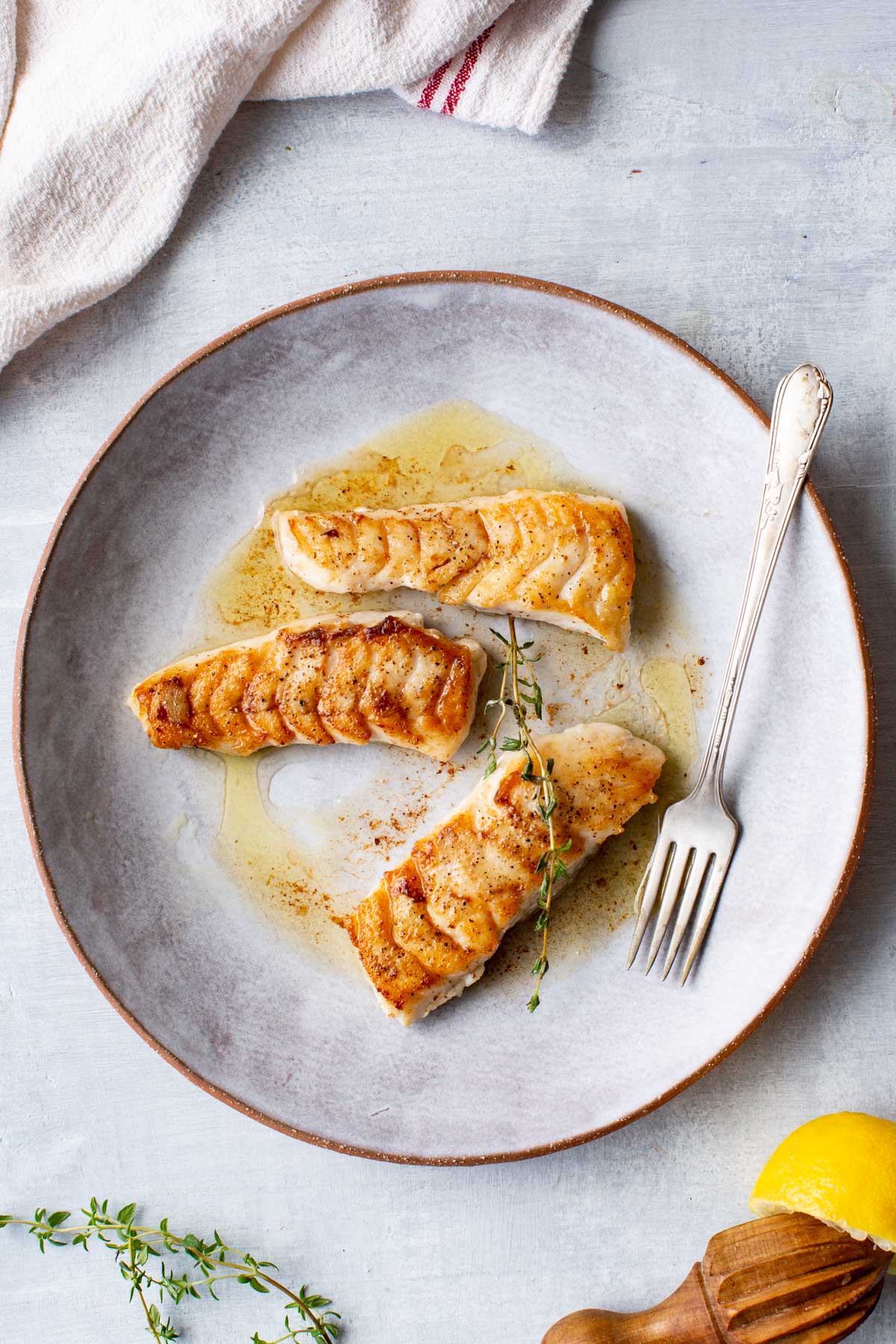 Three seared tilefish fillets plated with pan sauce and garnished with fresh thyme on a gray ceramic plate with a fork.