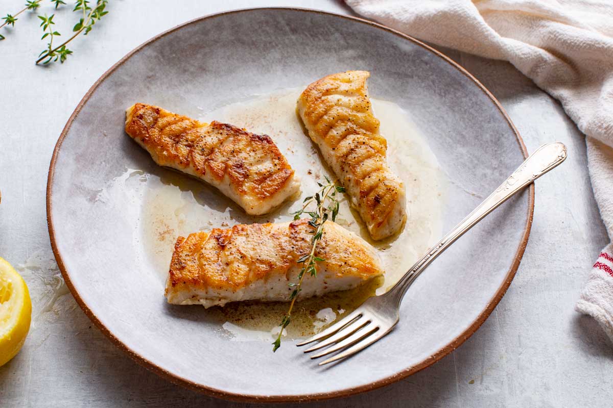 A side view of golden seared tilefish fillets with pan sauce on a gray plate, served with a fork and lemon wedge.