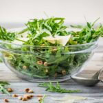 Glass bowl filled with baby arugula, shaved Parmigiano Reggiano, and toasted pine nuts on a rustic wood surface with lemon half and serving utensils.