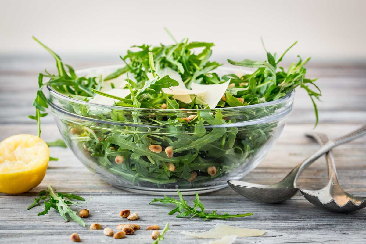 Glass bowl filled with baby arugula, shaved Parmigiano Reggiano, and toasted pine nuts on a rustic wood surface with lemon half and serving utensils.