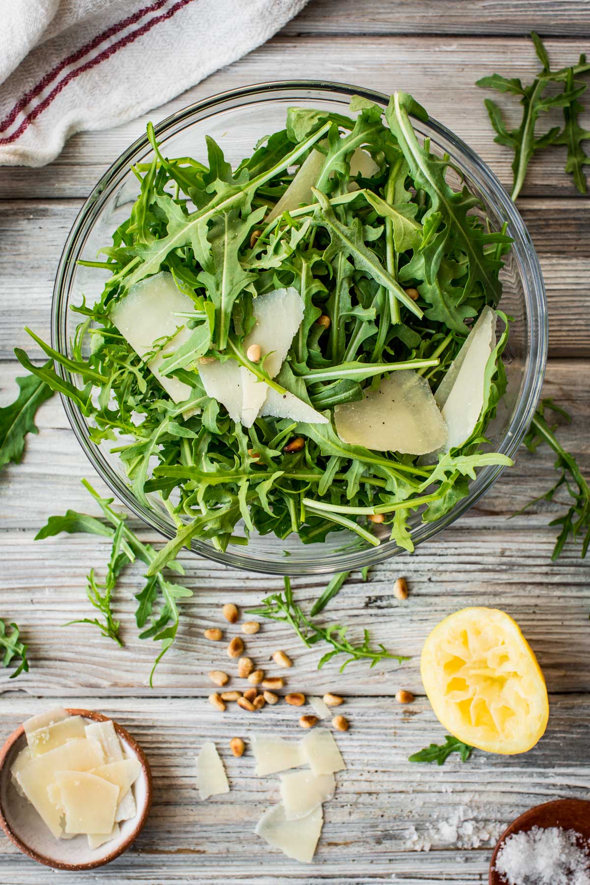 Overhead view of arugula salad with pine nuts and shaved parmesan in a glass bowl, surrounded by scattered cheese, arugula, lemon, and a pinch bowl of salt.