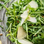 Close-up of arugula leaves, toasted pine nuts, and large shaved parmesan slices in a clear glass bowl.