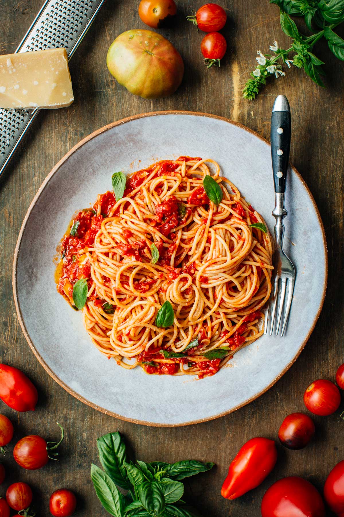 Overhead photo of spaghetti al pomodoro on a gray plate - A serving of spaghetti tossed with fresh tomato sauce and garnished with basil leaves, surrounded by fresh tomatoes, parmesan cheese, and basil on a rustic wooden table.