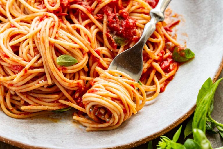 Angled close-up of spaghetti with fork – A fork twirls spaghetti coated in bright red pomodoro sauce, with bits of basil and parmesan visible on a shallow gray plate.