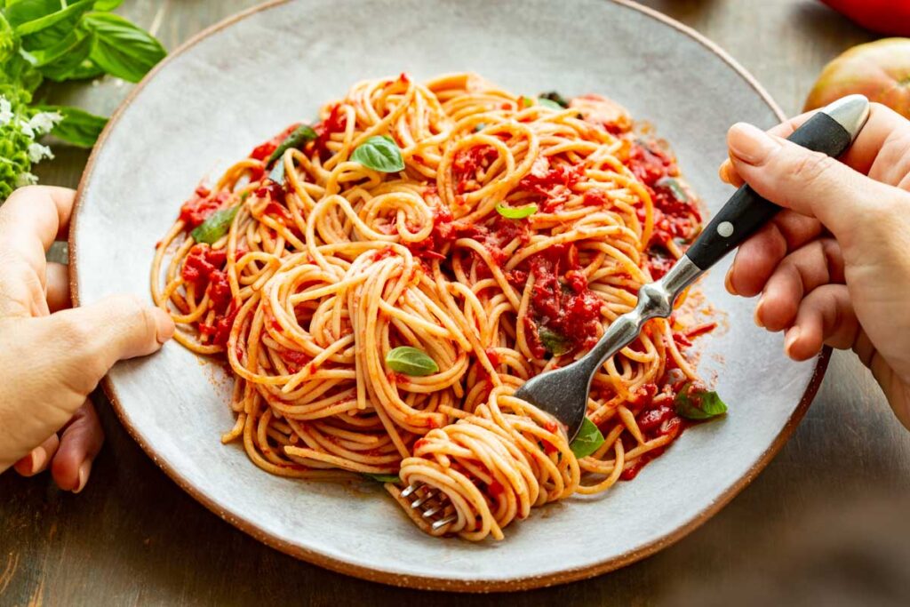 Hands holding a plate of spaghetti al pomodoro - A person twirls spaghetti with a fork while holding a plate full of pasta in fresh tomato sauce with basil leaves.