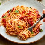 Hands holding a plate of spaghetti al pomodoro - A person twirls spaghetti with a fork while holding a plate full of pasta in fresh tomato sauce with basil leaves.