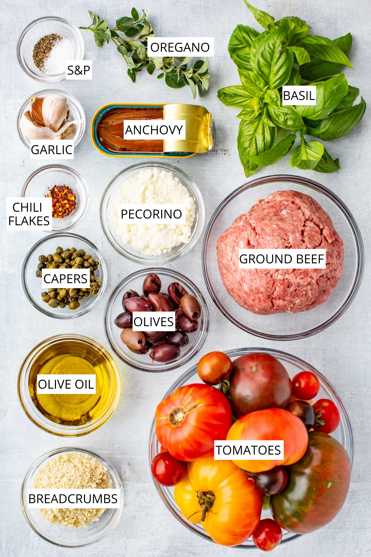 Overhead view of labeled ingredients for meatballs puttanesca including ground beef, anchovies, garlic, basil, oregano, tomatoes, olives, capers, breadcrumbs, olive oil, Pecorino, and chili flakes.