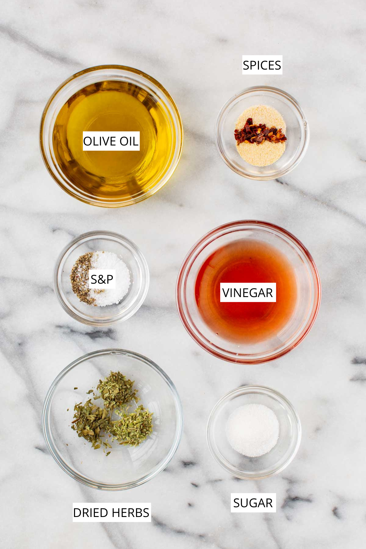 Overhead view of the basic ingredients for Italian dressing laid out on marble, including bowls of olive oil, red wine vinegar, dried oregano, garlic powder, onion powder, sugar, salt, black pepper, and red pepper flakes.