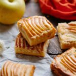 Two apple almond shortbread bars stacked on parchment paper, surrounded by whole apples and orange linen, highlighting the crisp edges and tender apple topping.