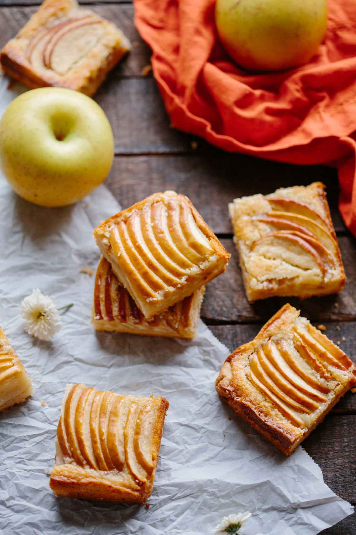 Overhead shot of apple almond shortbread bars with fanned apple slices on top, styled with whole apples, parchment paper, and scattered flowers.