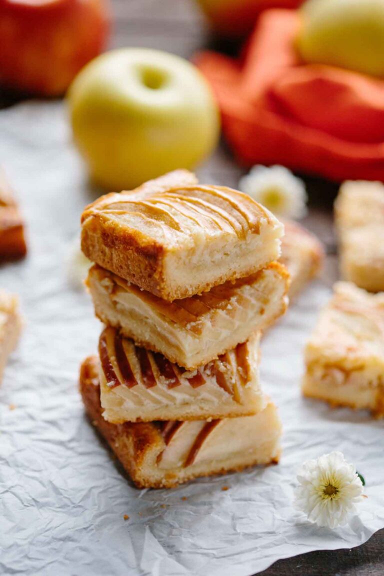 Stacked apple almond shortbread bars on parchment paper with whole apples in the background, showcasing the golden brown crust and sweet apple filling.