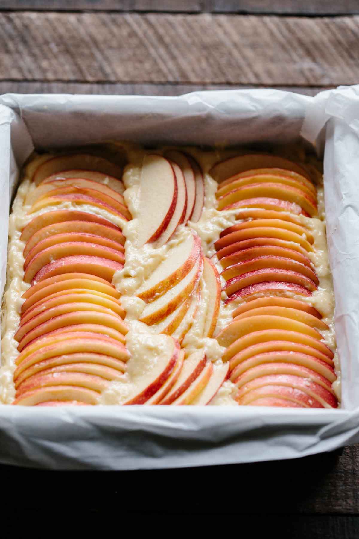 Unbaked apple almond shortbread bars in a parchment-lined pan, topped with rows of neatly arranged fresh apple slices ready for the oven.