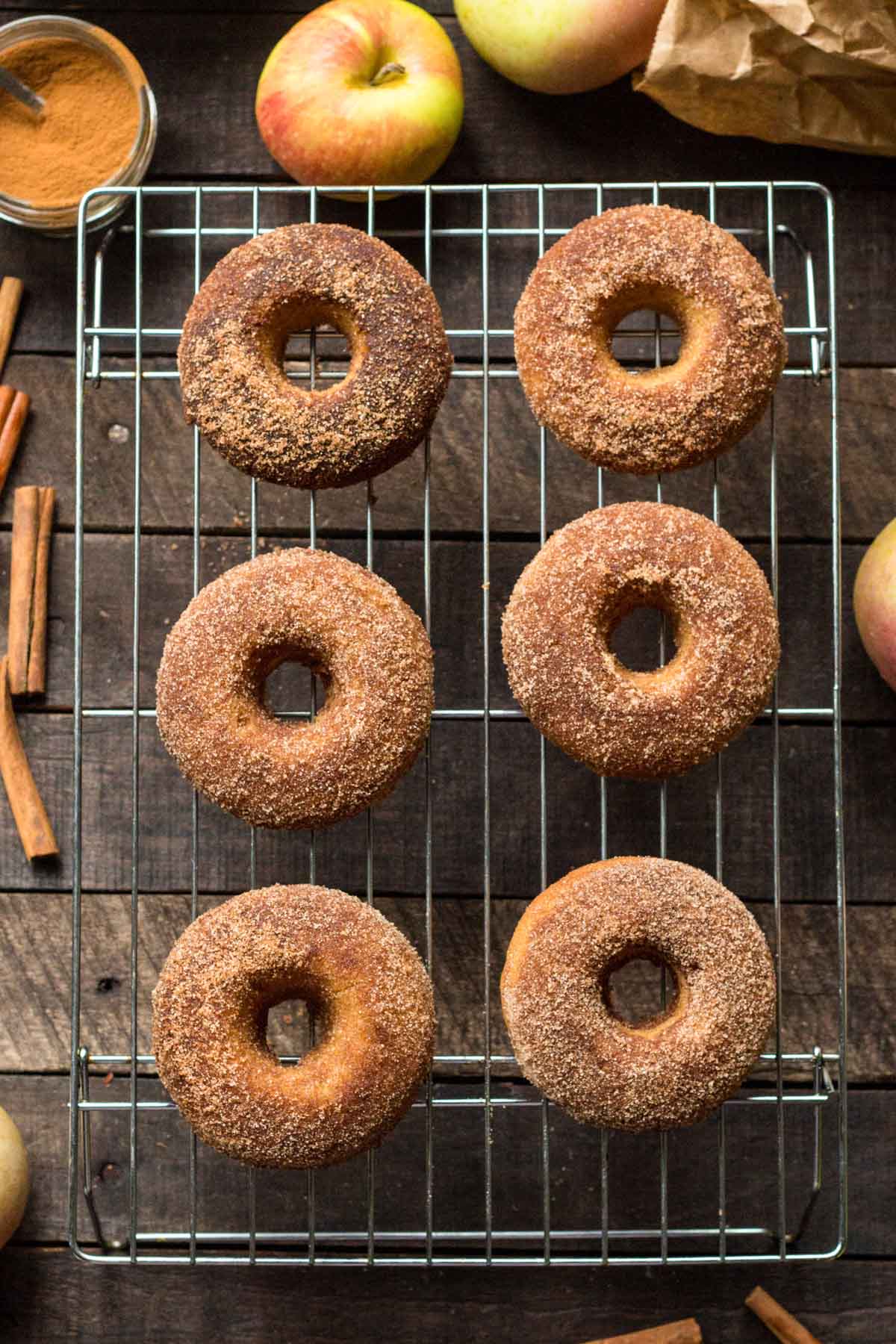Six baked apple cider donuts coated in cinnamon sugar resting on a wire cooling rack, with fresh apples, cinnamon sticks, and a jar of ground cinnamon scattered on a rustic wooden surface.
