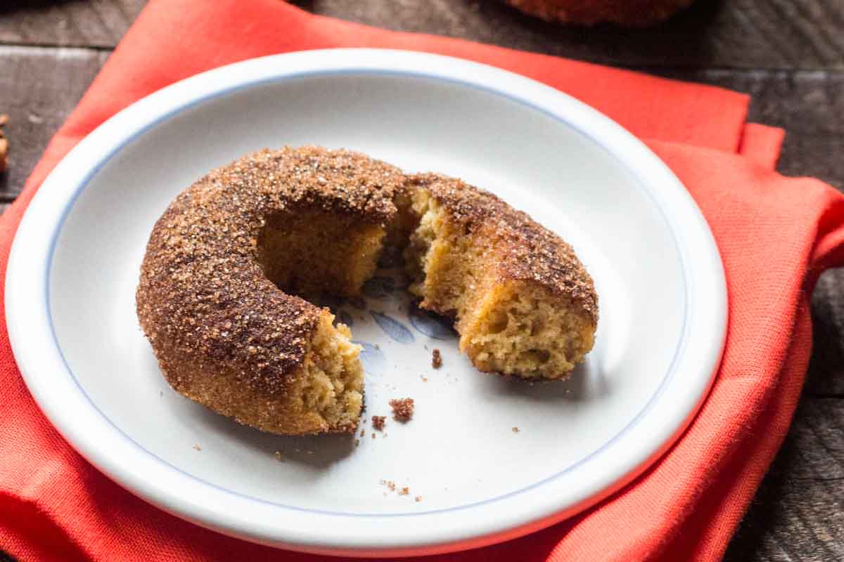 A single baked apple cider donut with a cinnamon sugar coating, broken in half to reveal its soft, cakey interior on a white plate with a red napkin.