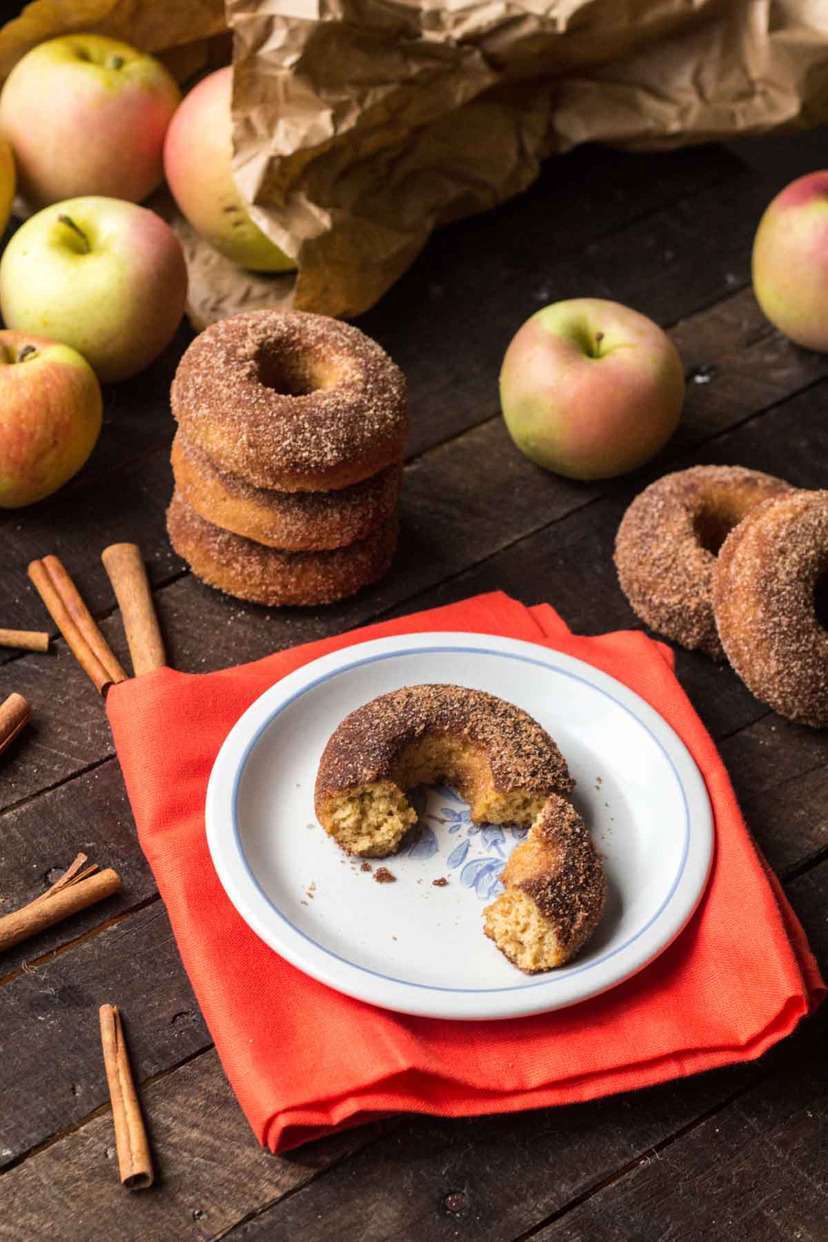 A cozy fall scene with baked apple cider donuts on a red napkin, broken open to show texture, surrounded by fresh apples, cinnamon sticks, and extra donuts on a rustic wooden table.