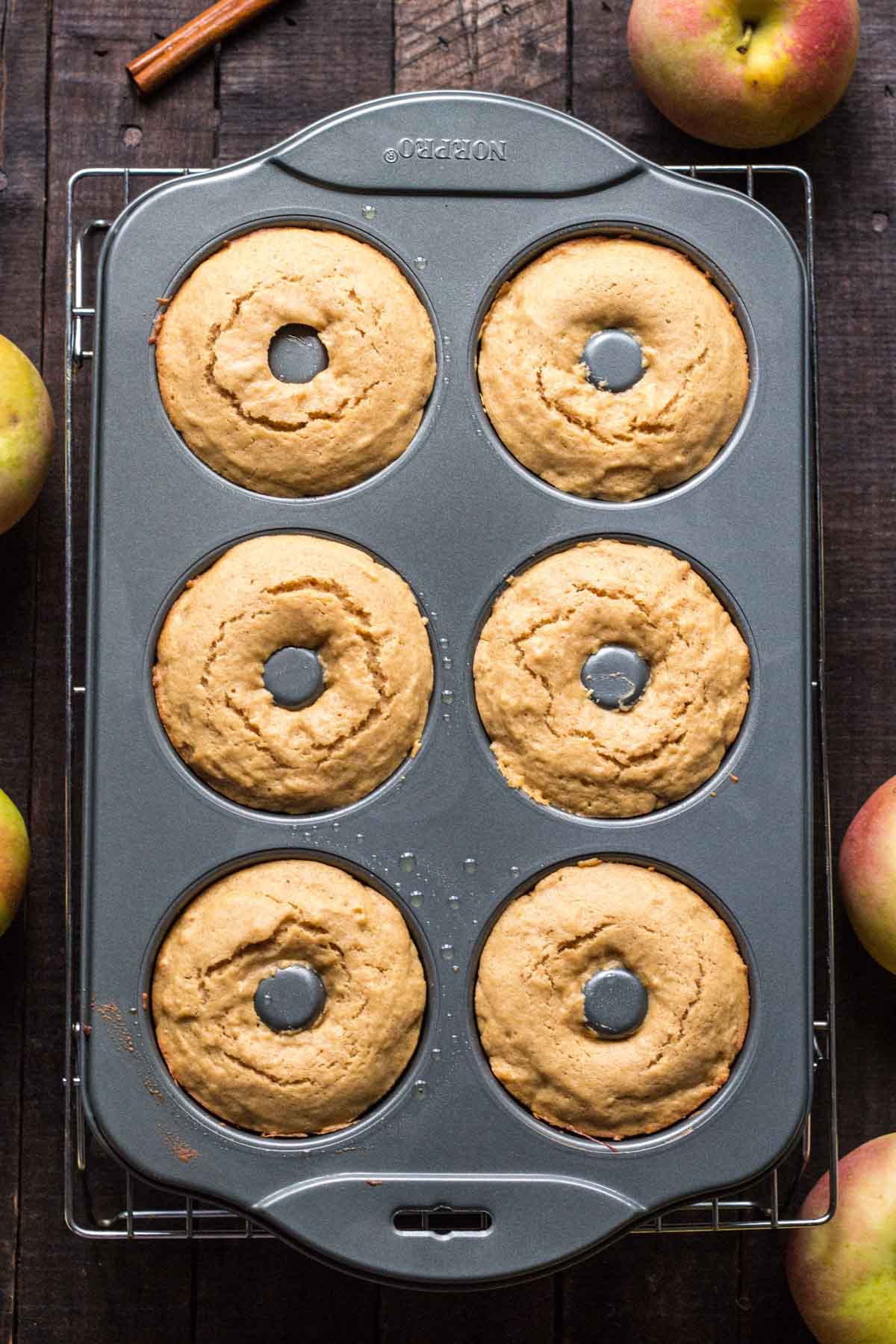 Freshly baked apple cider donuts still in a nonstick donut pan, golden brown and cooling, with apples scattered around on a wooden background.