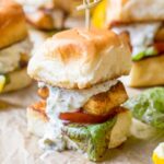 Golden brown fish slider on a toasted bun with tartar sauce dripping down the side, lettuce and tomato visible underneath.