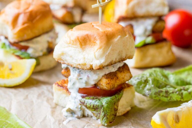 Golden brown fish slider on a toasted bun with tartar sauce dripping down the side, lettuce and tomato visible underneath.